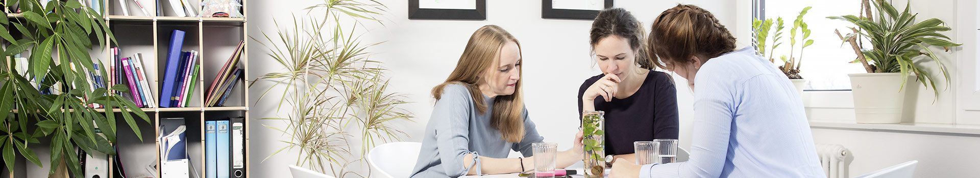3 Frauen sitzen in einem Raum mit weißem Bücherregal an einem weißen Tisch und lesen konzentriert.
