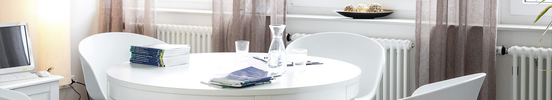 3 weiße Stühle stehen an einem weißen Tisch, auf dem ein Stapel mit Fachliteratur, 2 Gläser und eine Wasserkaraffe zu sehen ist.