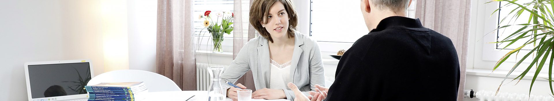 An einem weißen Tisch sitzt eine Psychotherapeutin mit Papier und einem Stift in der Hand und hört einer männlichen gestikulierenden Person aufmerksam zu.